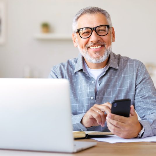 Handsome smiling senior man wearing glasses using mobile phone while sitting at his cozy workplace with laptop at home, retired male chatting with friends in social media, typing on smartphone